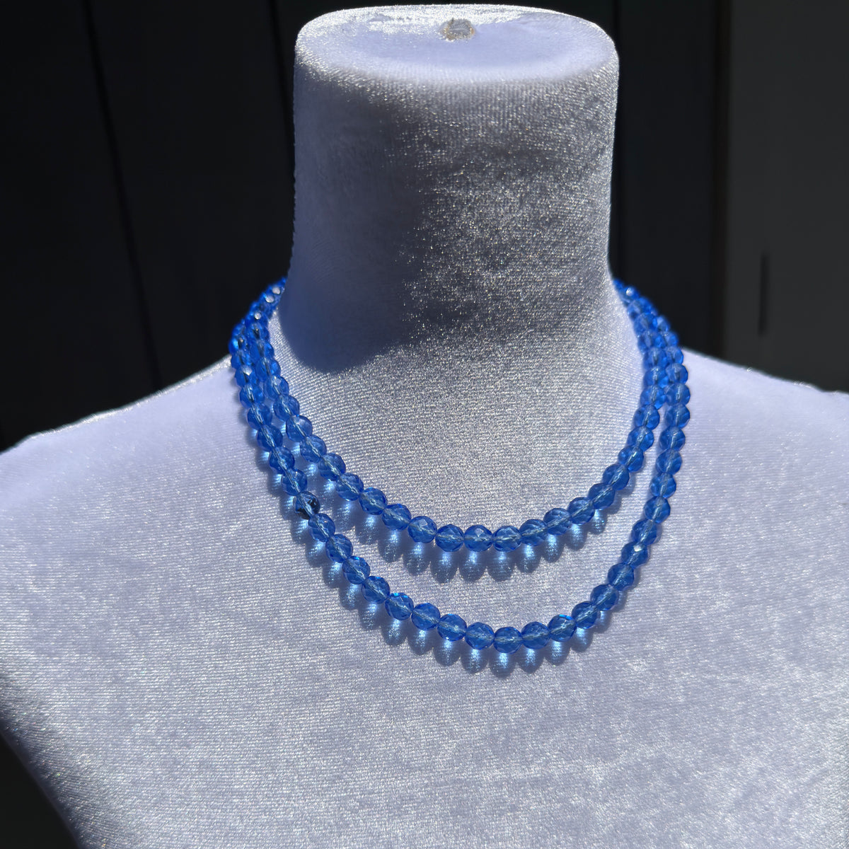 A double-strand necklace made of faceted blue beads, displayed on a light gray fabric jewelry bust.
